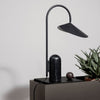 Sleek black lamp with a curved arm and geometric shade, set against a neutral background with green plant accents.