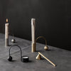 Sleek metal candle holders in gold, silver, and black with elegant curves on a gray tabletop against a dark backdrop.
