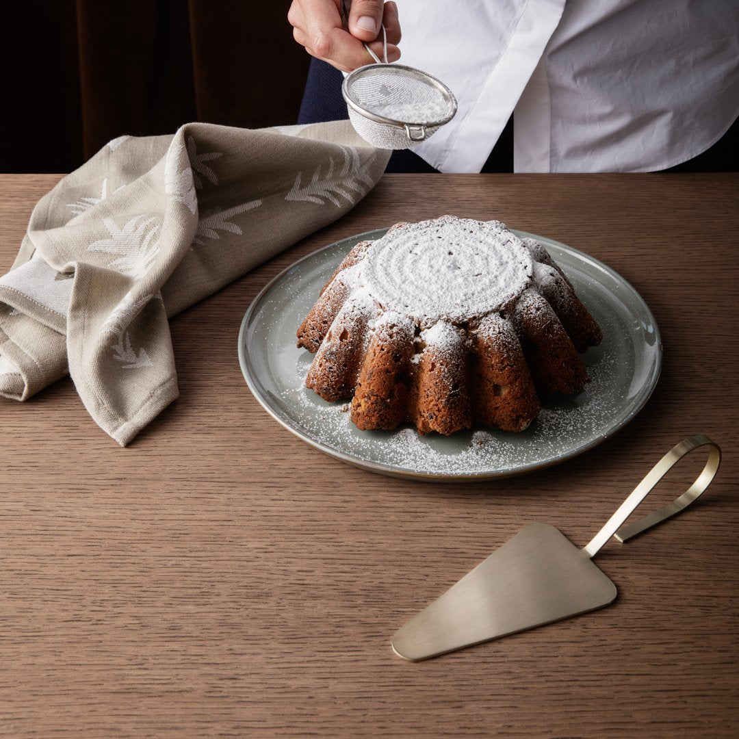 Brown bundt cake with powdered sugar on a gray plate, surrounded by a neutral-toned linen napkin and gold serving spatula.