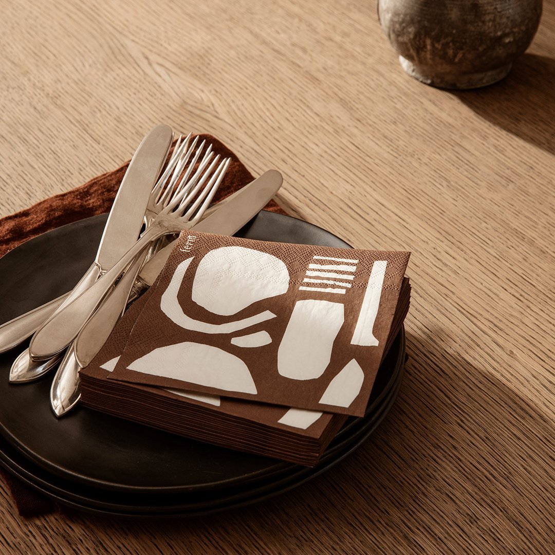 Brown napkins with white abstract patterns on a black plate, featuring silver cutlery, styled on a woven tablecloth.