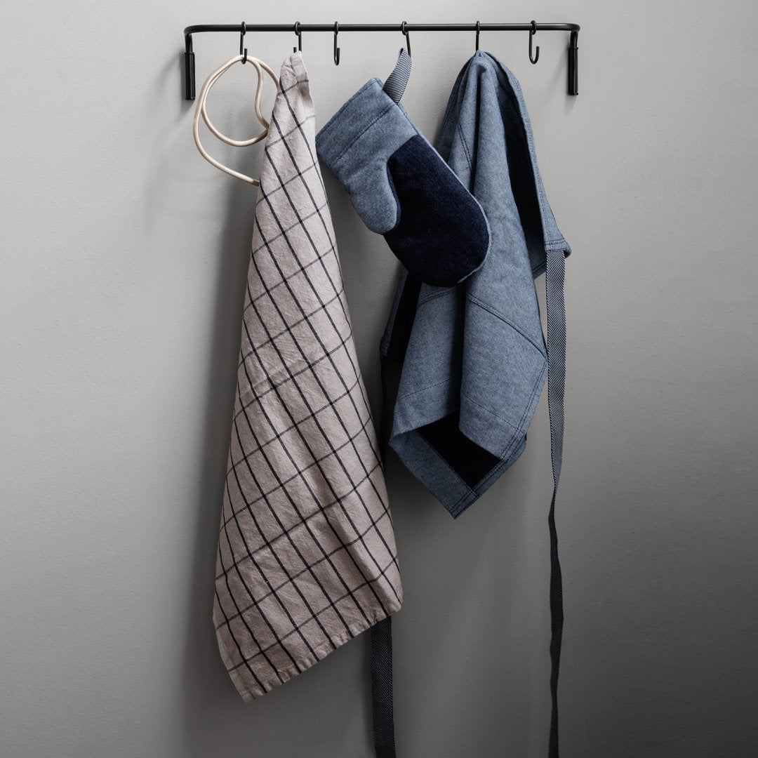 Gray apron with black grid pattern, blue kitchen towels, and oven mitt hanging on a wall hook.