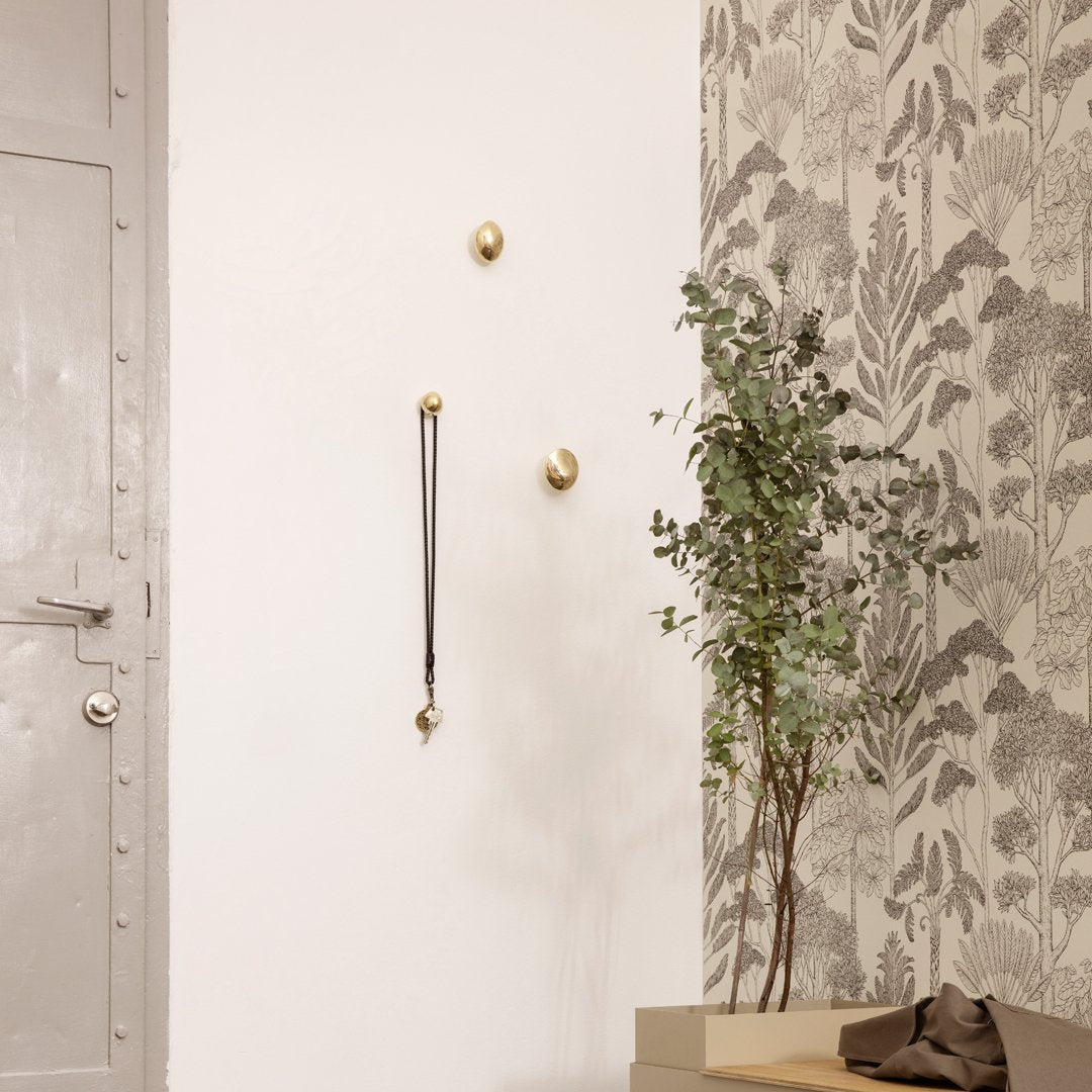 Brass wall hooks against a textured floral wallpaper in gray and white, with a green potted plant.