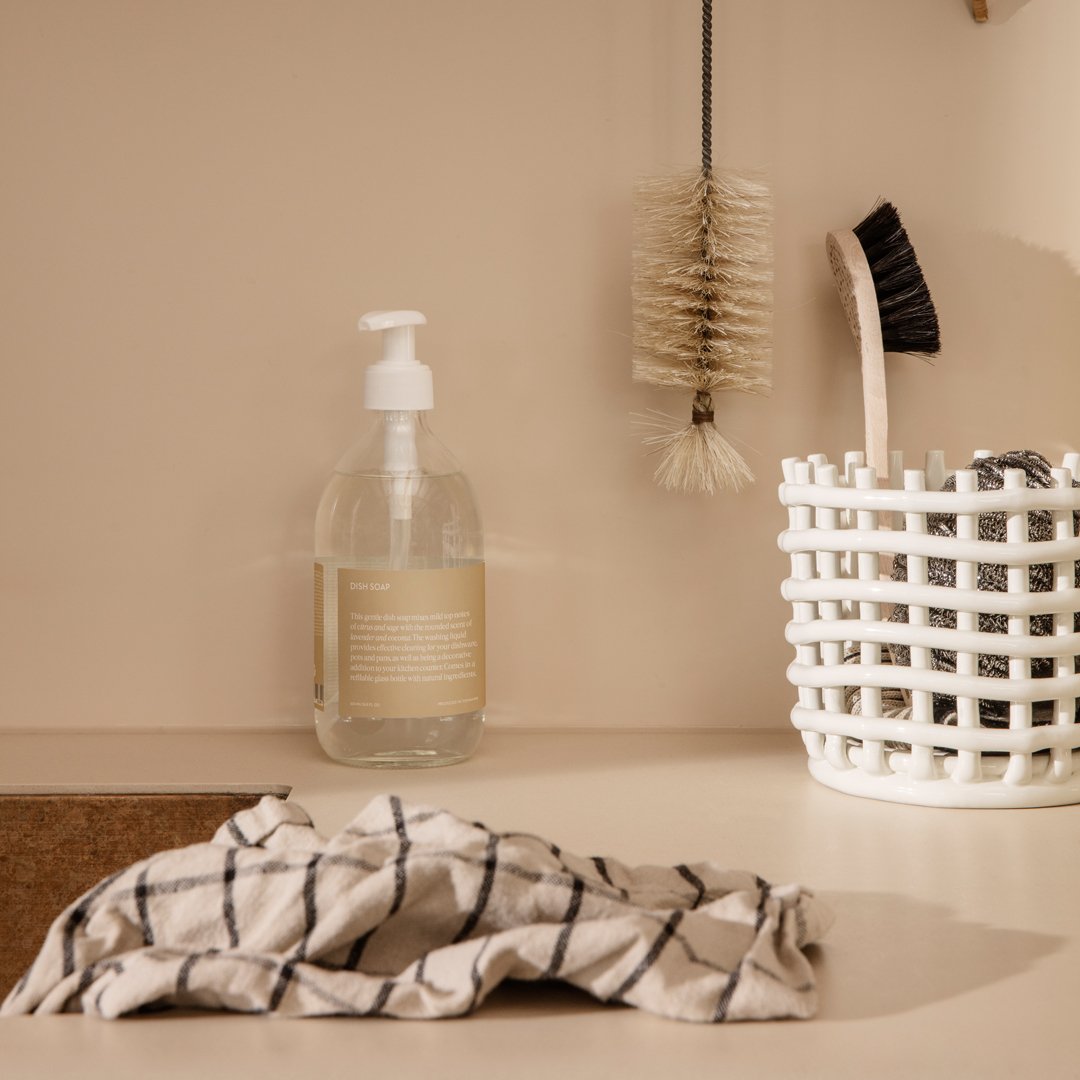 Clear glass soap dispenser, beige cloth with black grid pattern, white woven basket, and neutral background tones.