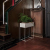 White planter with metal stand, featuring a lush green plant, set against dark wooden tones and red tile flooring.