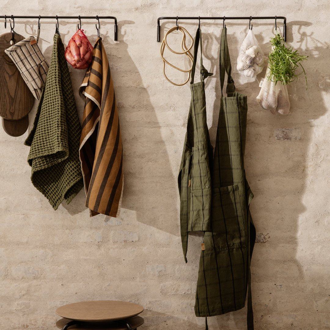 "Assorted aprons and kitchen towels in green, brown, and striped patterns, hanging against a textured wall."