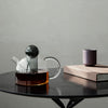 Clear glass teapot with black lid, amber tea, and a minimalist purple cup on a sleek black table.