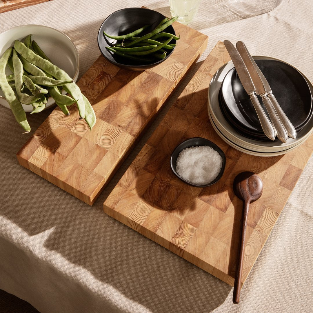 Wooden cutting boards with a warm checkerboard pattern, black ceramic bowls, and natural green vegetables on a light tablecloth.