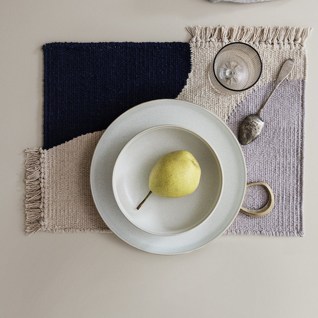 Multicolored woven placemat featuring navy, beige, and lavender sections with fringe detailing. Ideal for modern dining.