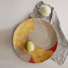 Round ceramic plate featuring abstract patterns in warm yellow, earthy tones, and a bold red accent.