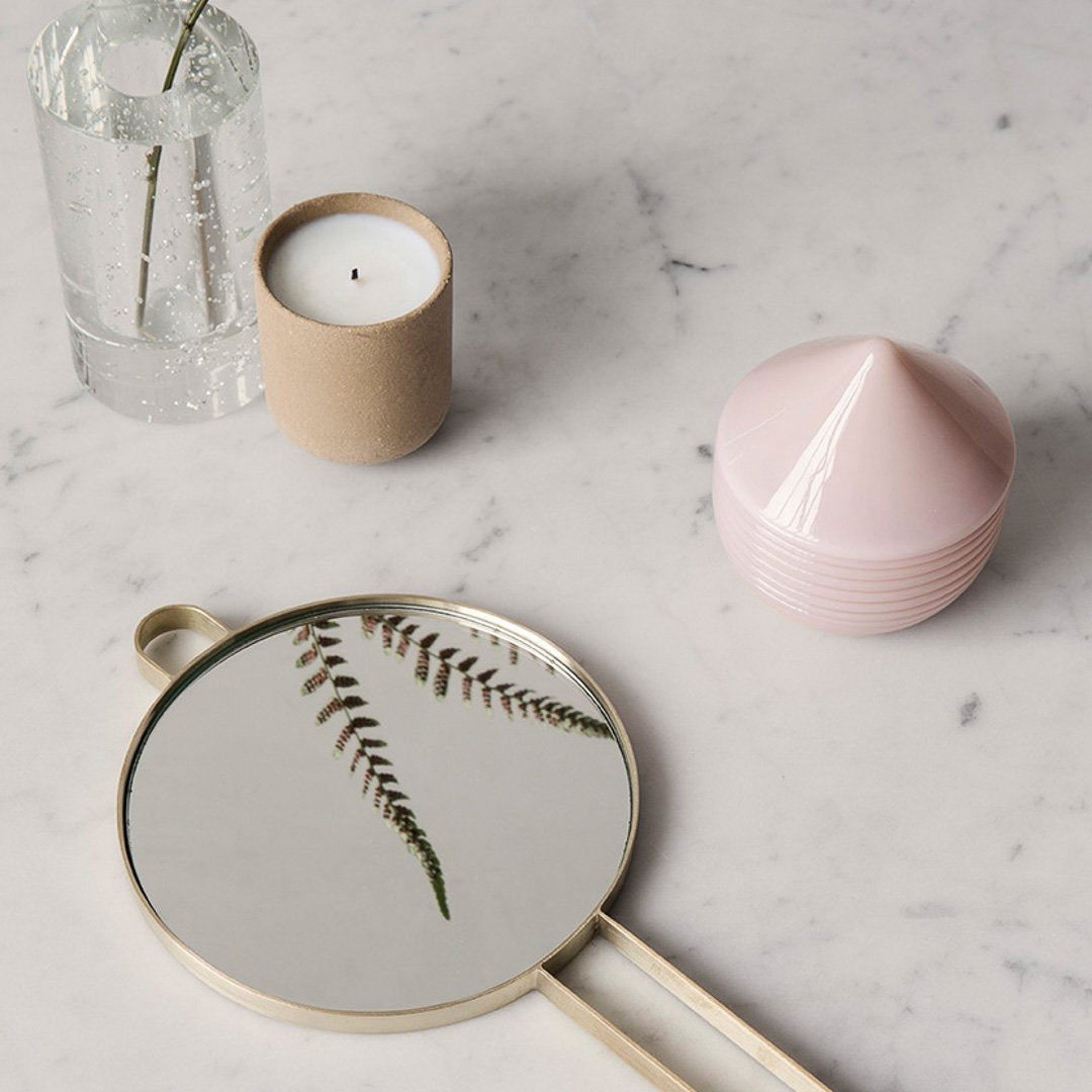 Round mirror with gold trim reflecting green fern, soft pink and beige woven storage, clear glass vase, white candle.