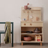 "Wooden kitchen playset in light tones with pink and earthy colored accessories on open shelves and drawers."
