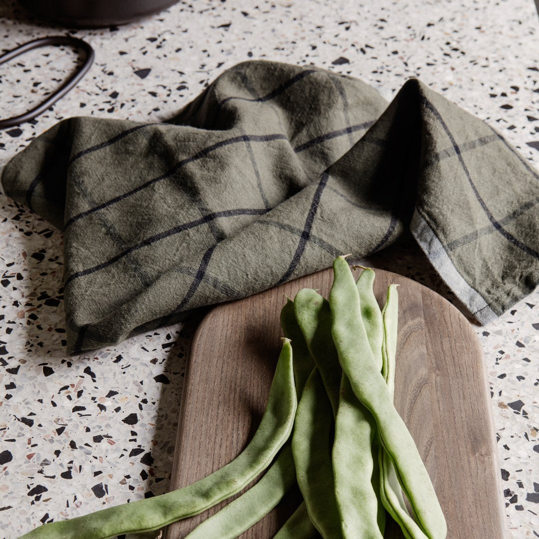 Olive green dish towel with a black grid pattern, resting on a wooden board with green beans.