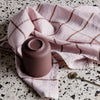 Pink linen with brown grid pattern and mauve ceramic cup on speckled surface. Soft, modern color palette.