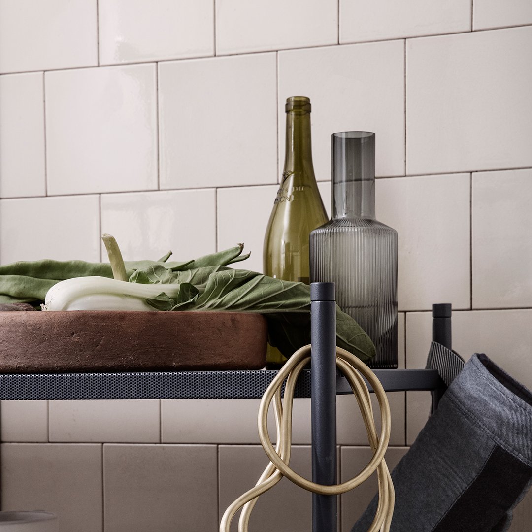 Green and grey glass bottles, earthy tones of the bowl, and vibrant leafy greens on a black shelf against a pale backdrop.