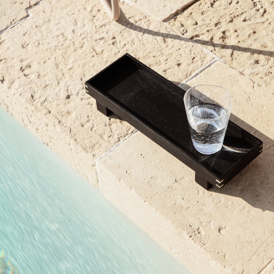 Black rectangular tray with smooth surface, positioned next to a turquoise pool, holding a clear glass of water.