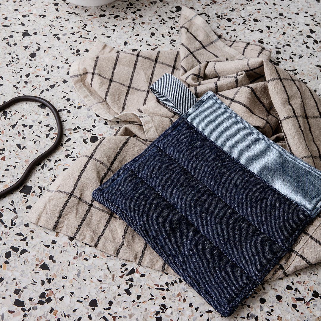 Denim pot holder in navy and light blue on a beige checkered towel, set against a speckled countertop.