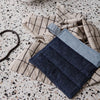 Denim pot holder in navy and light blue on a beige checkered towel, set against a speckled countertop.