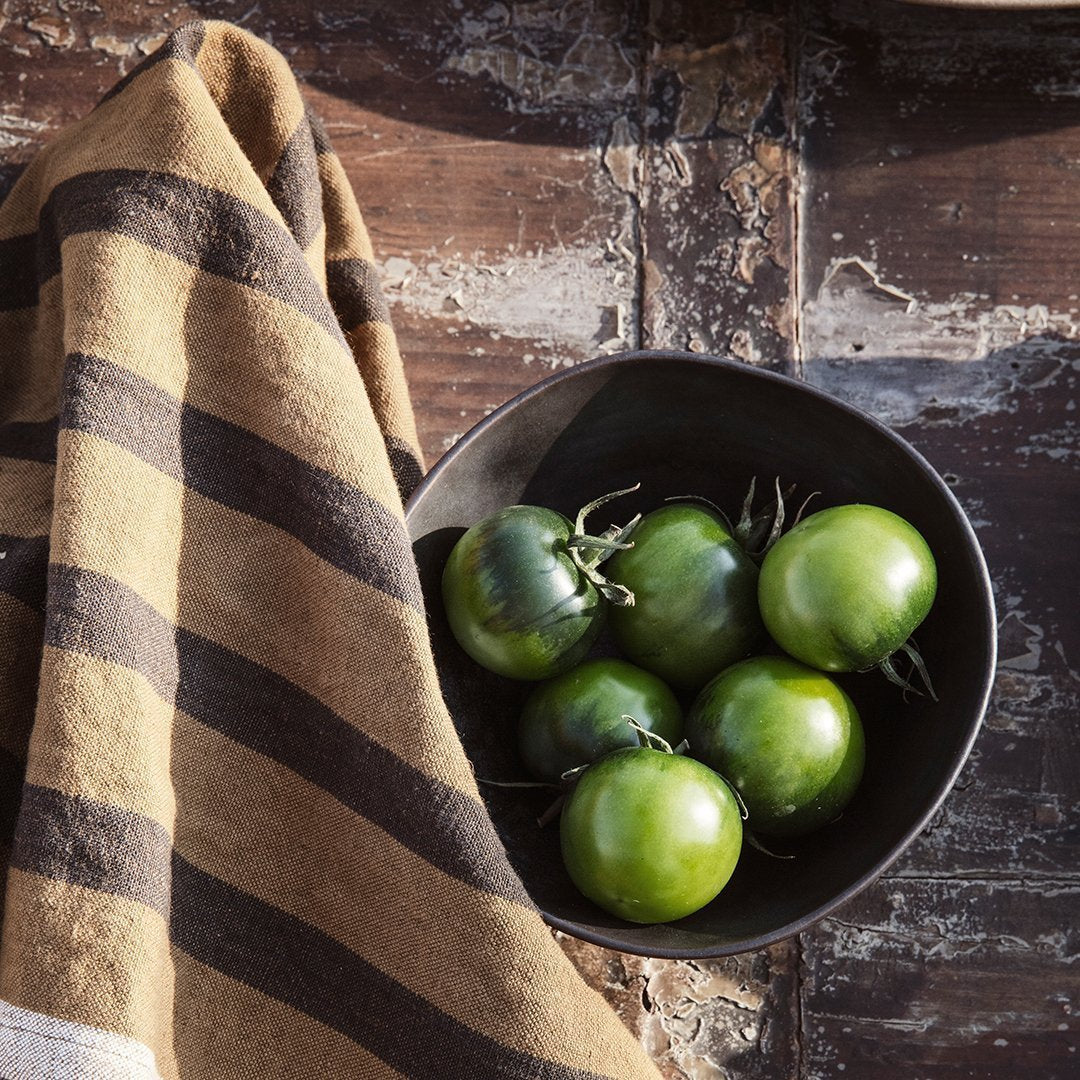 Fresh green tomatoes in a black bowl, accompanied by a striped tan and brown towel against a rustic wood background.