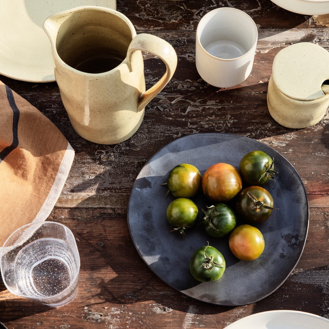 "Round black plate with green and orange tomatoes, rustic ceramic pitcher, and assorted dishware on a wooden table."