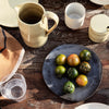 Rustic table setting with earthy tones, green and yellow tomatoes on a dark plate, surrounded by minimalist ceramics.
