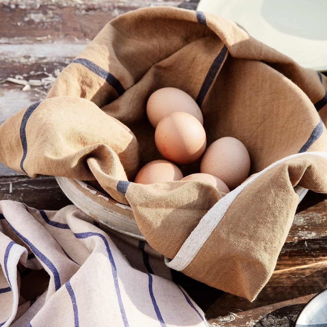 Brown linen cloth with navy stripes, draped over a bowl of eggs, on a rustic wooden surface.