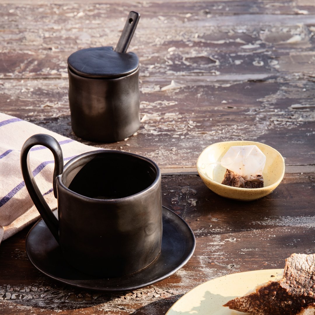 Black ceramic coffee cup and lid, sleek design on rustic wooden table with a yellow dish and striped cloth.