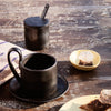 Black ceramic coffee cup and lid, sleek design on rustic wooden table with a yellow dish and striped cloth.