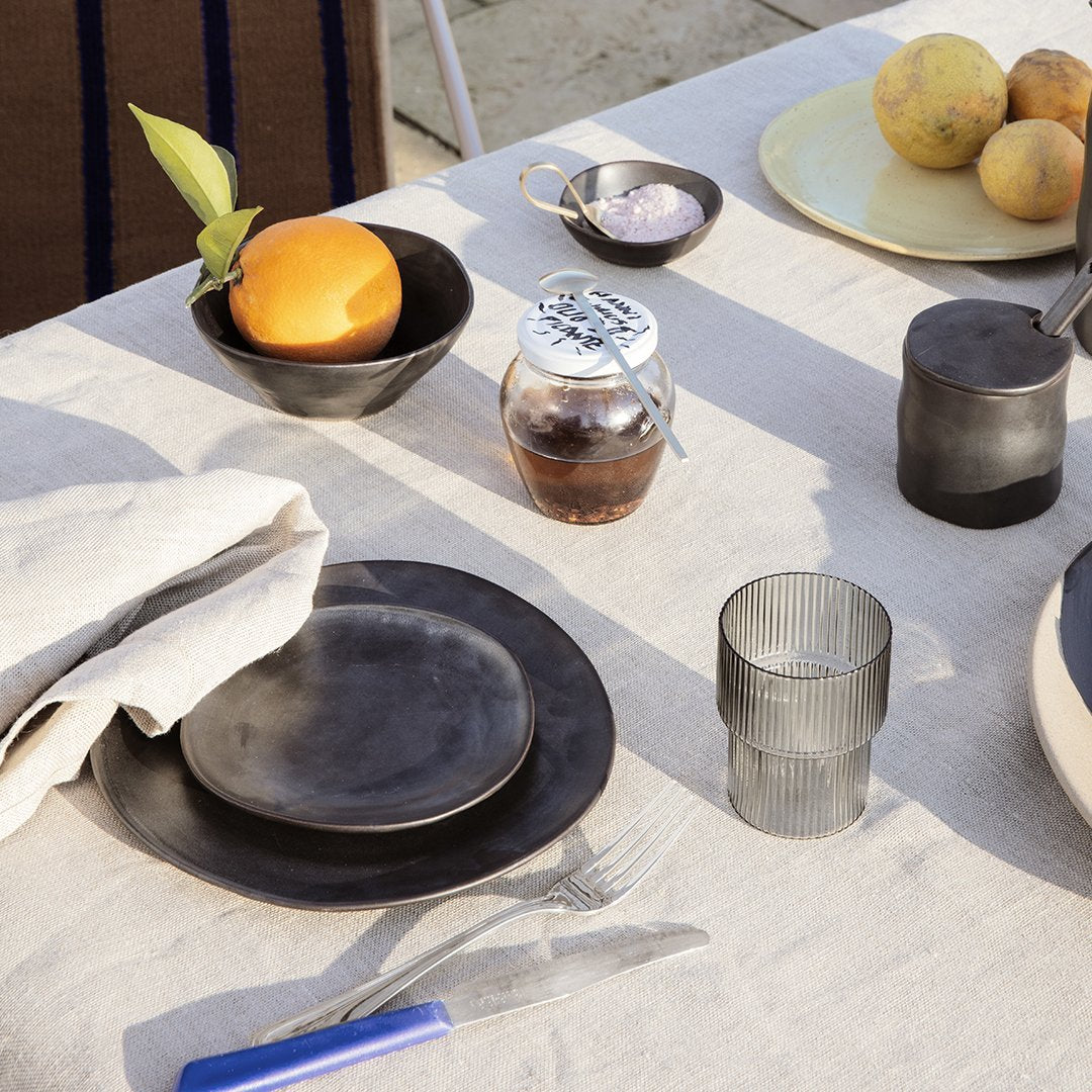 Black ceramic tableware with textured glass; neutral-toned tablecloth and colorful fruits add vibrant accents.