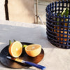 Blue basket, black plate, cut orange, green leaves, and a blue-handled knife on a beige tablecloth.