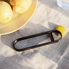 Black oval kitchen tool on a light beige linen surface with lemons and a yellow bottle cap.