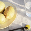 "Yellow lemons on a soft beige plate with a clear glass of water and dark utensils on a textured, light linen backdrop."