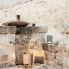 Decorative earthy tones and textures, featuring a woven pillow, ceramic pots, and greenery against a stone backdrop.