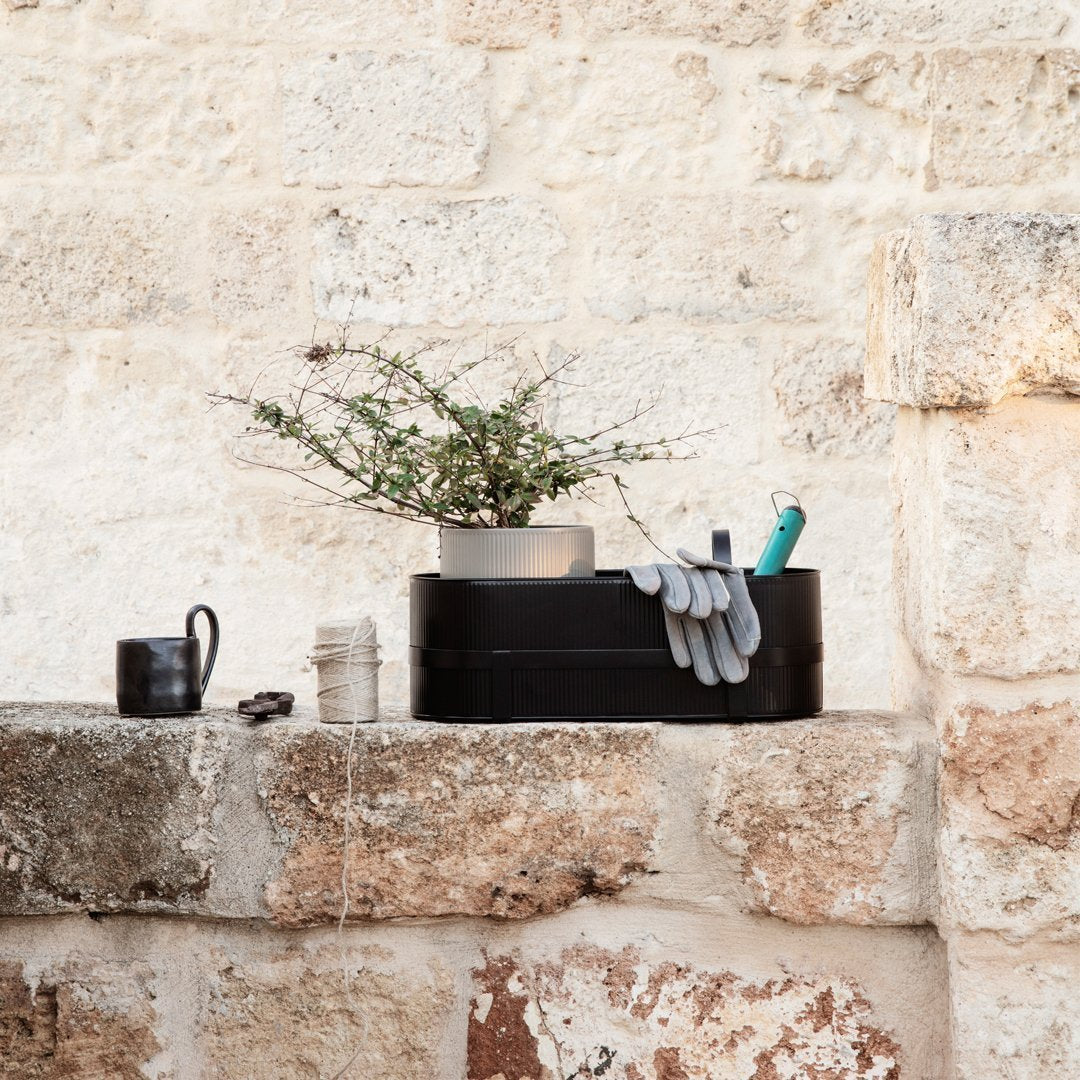 Black storage box with gray gloves and green tools, set against a textured beige stone wall, featuring a potted plant.