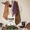 "Set of kitchen towels in earthy tones with stripes and checks, hanging above rustic kitchen items."