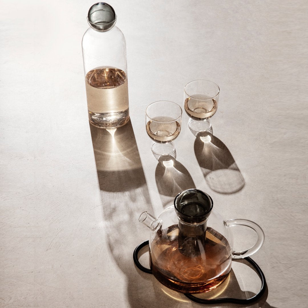 Clear glass carafe and cups, featuring warm amber liquid, on a light surface with soft shadows. Elegant modern design.