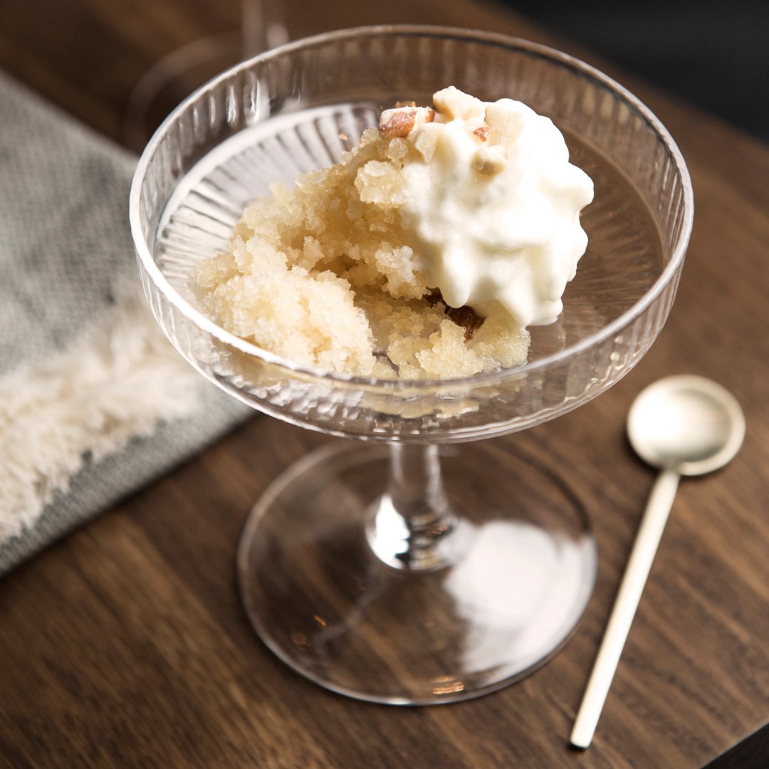 Clear glass dessert bowl with textured rim, filled with light yellow sorbet and white cream on a dark wooden surface.