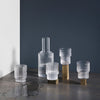 Frosted glass drinkware set with clear and gold accents against a dark gray background. Modern, minimalist design.