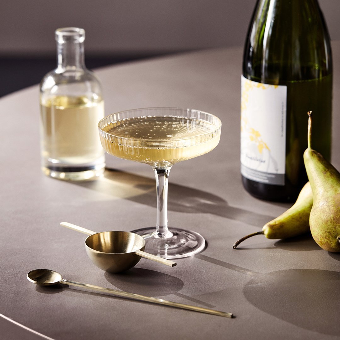 Elegant champagne cocktail in a textured glass, paired with gold utensils and fresh green pears on a gray surface.