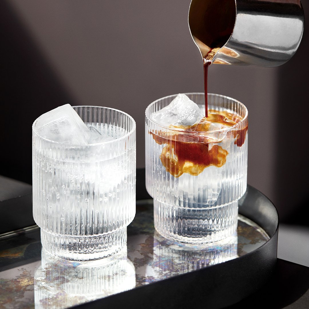 Clear ribbed glasses with ice, featuring contrasting dark liquid pour, set on a reflective tray.
