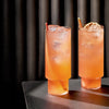 "Two textured orange cocktail glasses on a dark surface, showcasing a vibrant gradient with ice and garnish."