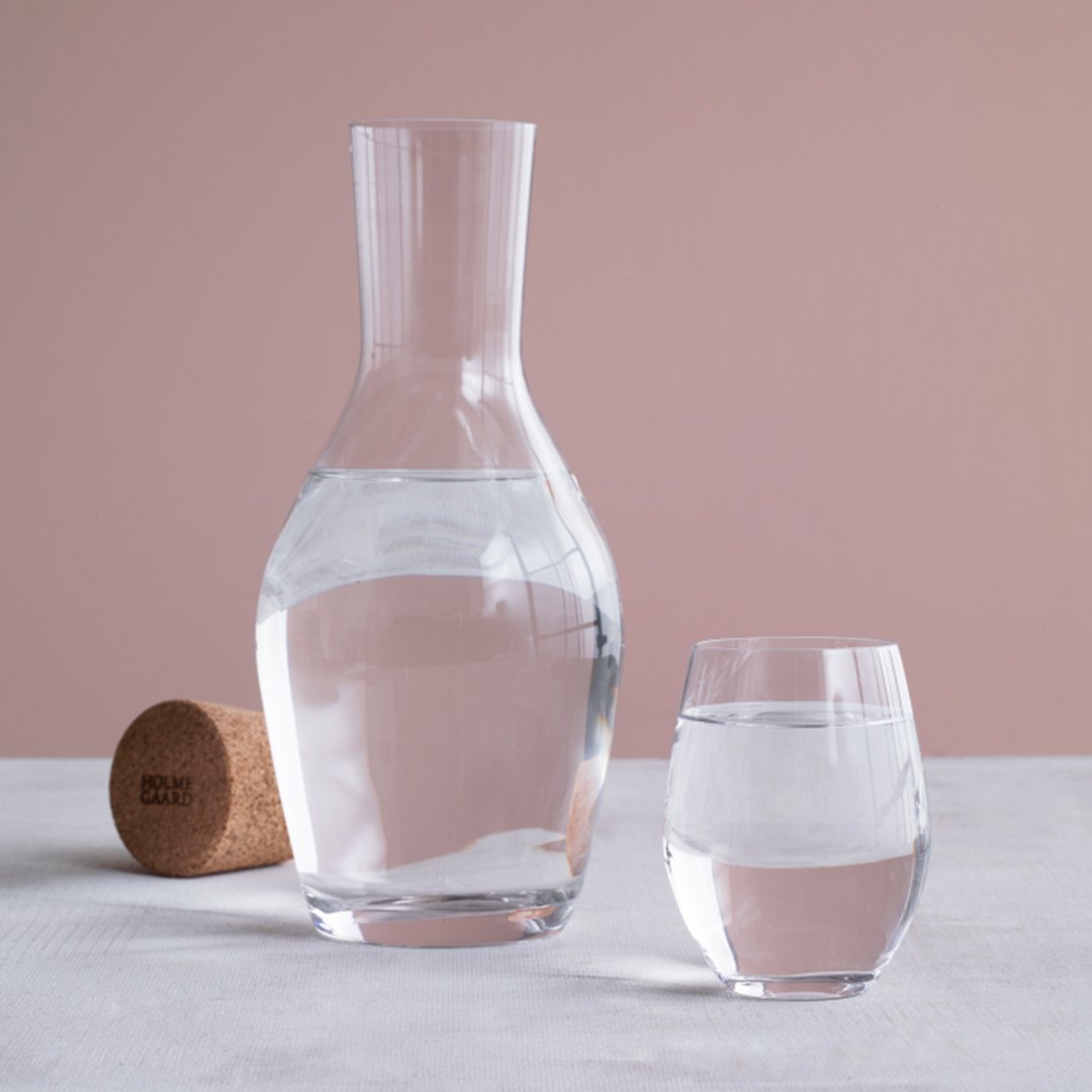 Clear glass water carafe and tumbler on a pale pink background, featuring smooth, sleek lines and subtle reflections.
