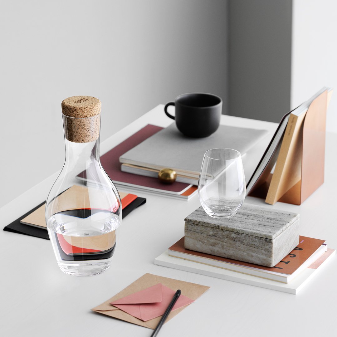 Clear glass carafe with cork stopper, modern patterns in shades of beige, copper, and muted red on a minimalist desk.