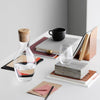 Clear glass carafe with cork stopper, modern patterns in shades of beige, copper, and muted red on a minimalist desk.