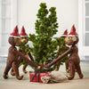 Wooden monkey figurines in red hats surround a green Christmas tree with a red gift and burlap sack.