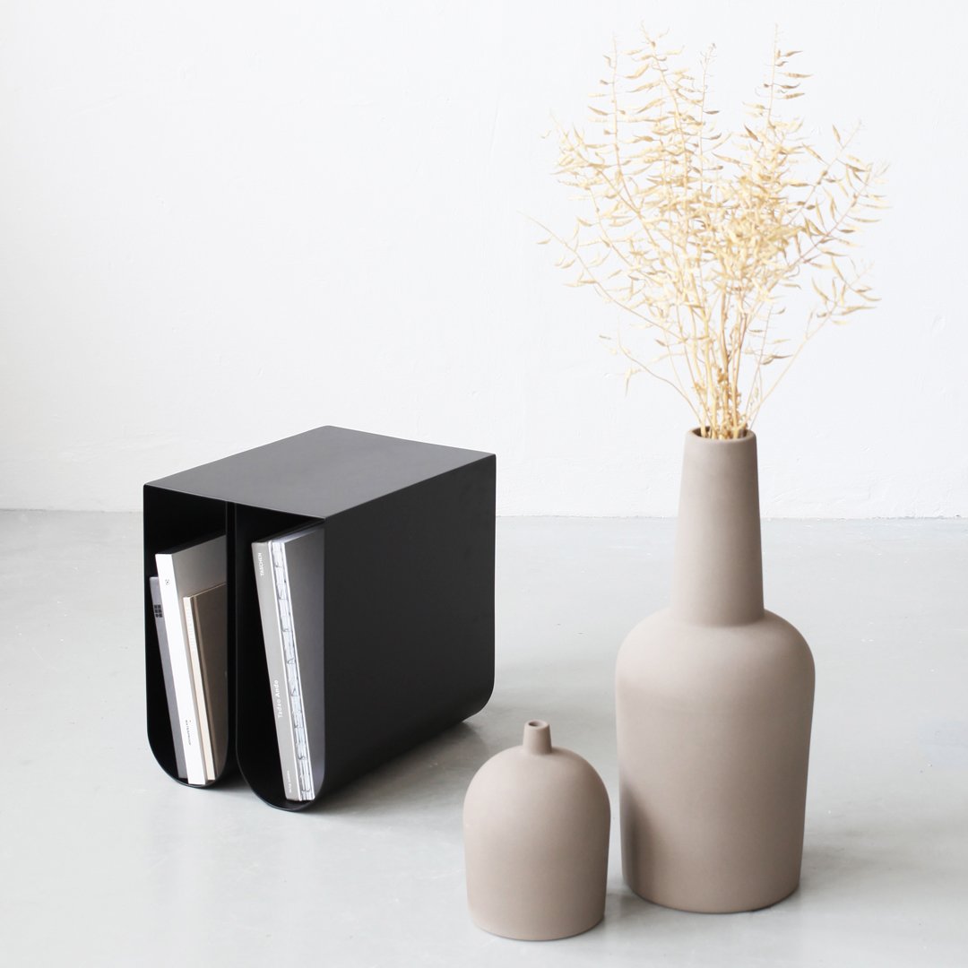 Black metal magazine holder with beige vases and dried flowers, featuring smooth matte finishes and modern shapes.