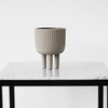 Gray ribbed ceramic planter with three legs, displayed on a marble table surface. Minimalist design.