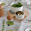 "White bowls with greens and olives on a light wooden tray, set against a neutral tablecloth."