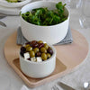 White ceramic bowls on a wooden platter, featuring greens in the larger bowl and olives with feta in the smaller bowl.
