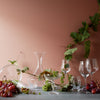 "Elegant glassware on a soft pink backdrop, adorned with green vines and clusters of purple and green grapes."