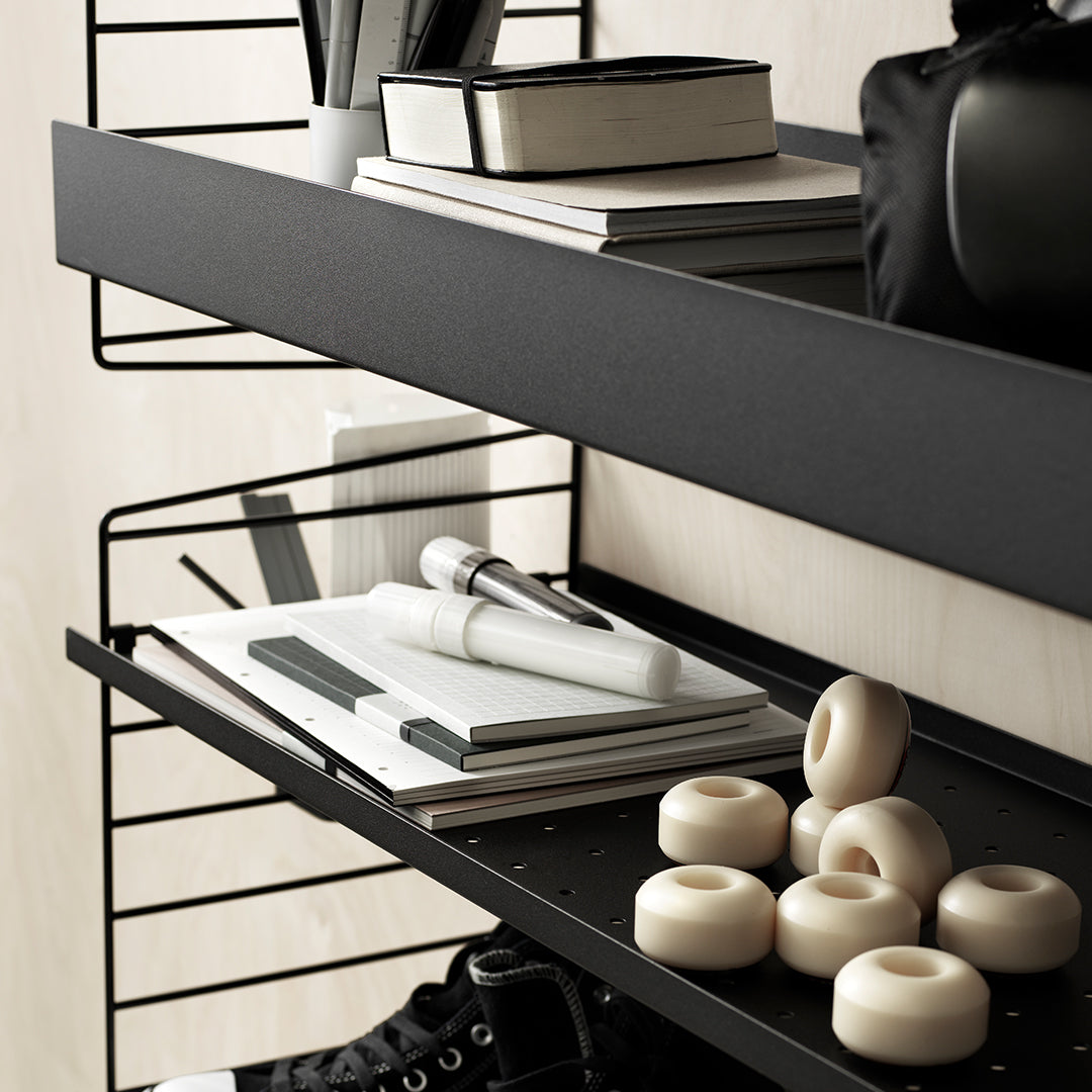 Black metal shelving with a minimalist pattern, featuring white notebooks and beige decorative items.
